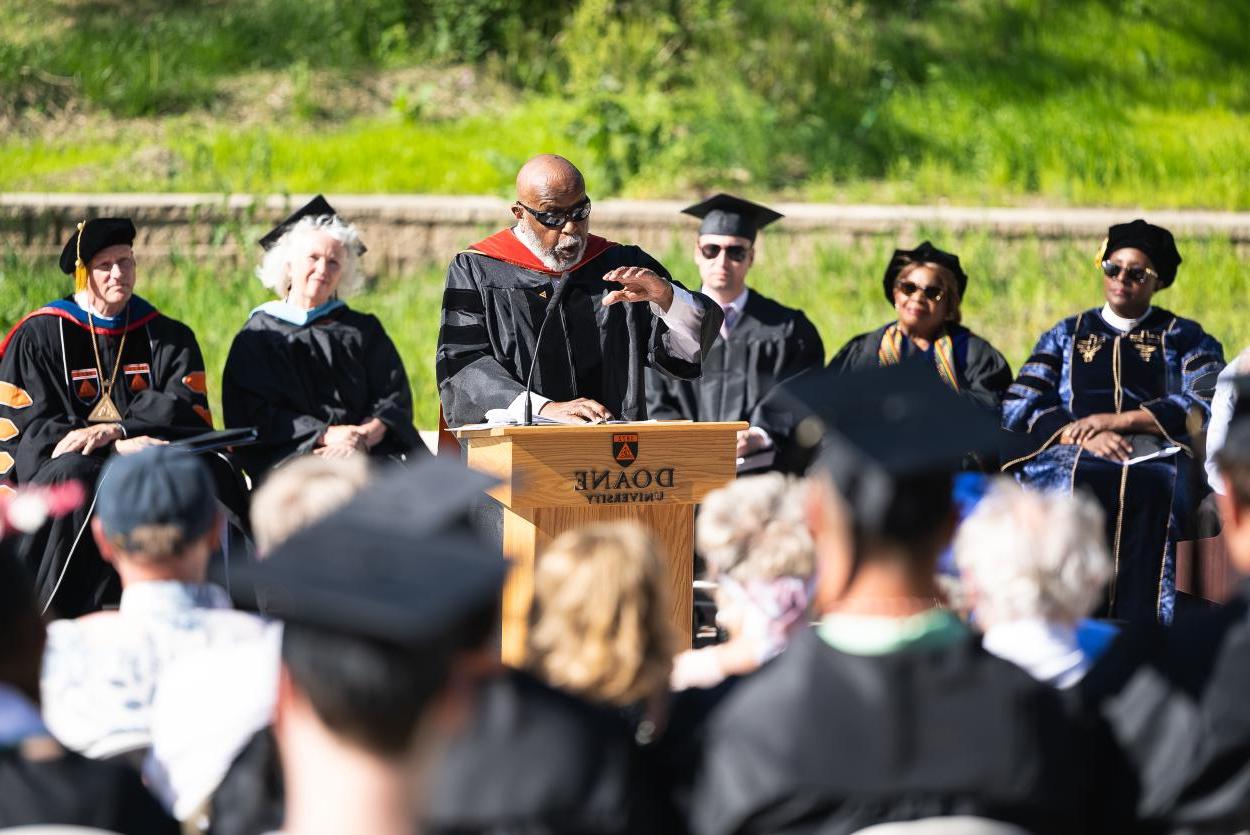 A Black man in an academic robe gestures over a podium toward an audience wearing black robes and graduation caps. Behind him are five people, also in academic robes.